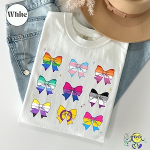May include: White t-shirt featuring colorful bow designs representing various LGBTQ+ pride flags. The bows are arranged in a grid pattern. The shirt is displayed on a denim background with a hat.