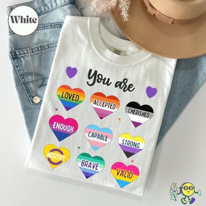May include: White t-shirt with the words "You are" above colourful heart graphics. Each heart features a word: Loved, Accepted, Cherished, Enough, Capable, Strong, Worthy, Brave, and Valid. The shirt is folded on a pair of blue jeans.