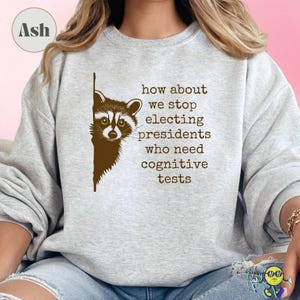 Ash gray sweatshirt worn by a model with a brown raccoon peeking from the left side. Text in typewriter-style font reads, "how about we stop electing presidents who need cognitive tests." The design is centered on the chest. The model is seated, wearing ripped light blue jeans.