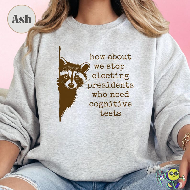 Ash gray sweatshirt worn by a model with a brown raccoon peeking from the left side. Text in typewriter-style font reads, "how about we stop electing presidents who need cognitive tests." The design is centered on the chest. The model is seated, wearing ripped light blue jeans.
