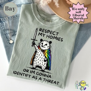 Peut inclure: Un t-shirt vert clair avec l'inscription "RESPECT MY HOMIES OR IM GONNA IDENTIFY AS A THREAT". Le motif représente un opossum en cape arc-en-ciel tenant une épée. Le t-shirt est présenté avec un jean bleu et un chapeau.