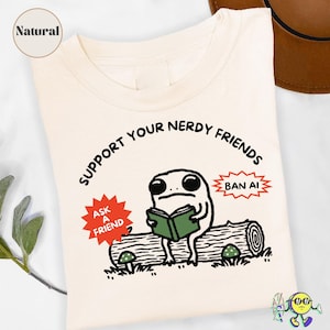 Könnte beinhalten: Ein naturfarbenes T-Shirt mit einer Grafik eines Frosches, der auf einem Baumstamm sitzt und ein grünes Buch liest. Der Text lautet "SUPPORT YOUR NERDY FRIENDS", "ASK A FRIEND" in einem roten Stern und "BAN AI" in einem roten Stern.