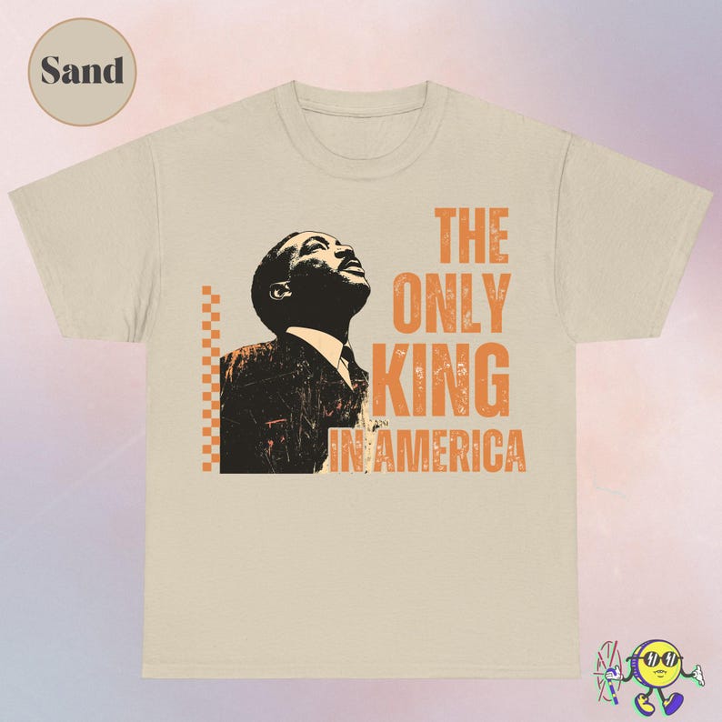 Sand-colored T-shirt with a graphic of a man in black and beige looking upward. To the right of the image, distressed orange text reads "THE ONLY KING IN AMERICA." A vertical orange and beige checkerboard pattern is to the left of the graphic. The fabric appears soft with a classic crew neck and short sleeves.