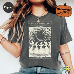 A model wearing a faded dark gray "Pepper" t-shirt with a vintage-style cream graphic of five witches holding hands in a circle beneath a crescent moon and stars, framed by bare tree branches. The text above the graphic reads "VIVE LA RÉSISTANCE" with moon phase symbols surrounding it. A Halloween pumpkin graphic with "Halloween drop" text appears in the top right corner.