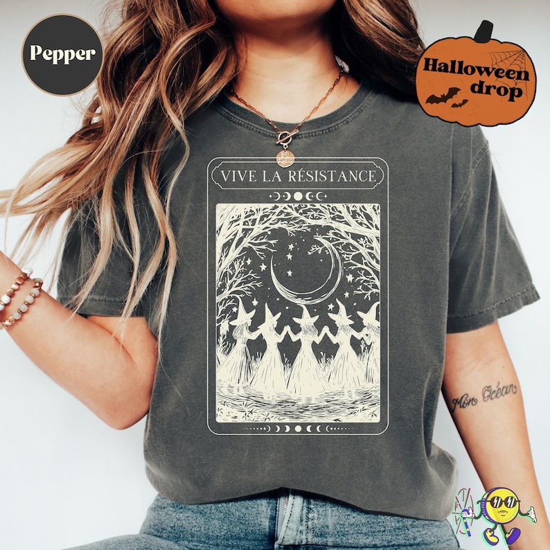 A model wearing a faded dark gray "Pepper" t-shirt with a vintage-style cream graphic of five witches holding hands in a circle beneath a crescent moon and stars, framed by bare tree branches. The text above the graphic reads "VIVE LA RÉSISTANCE" with moon phase symbols surrounding it. A Halloween pumpkin graphic with "Halloween drop" text appears in the top right corner.