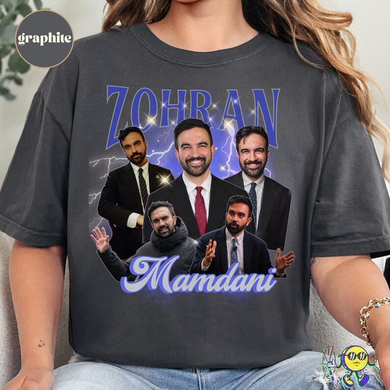 Graphite-colored t-shirt featuring a collage of five images of a man in various outfits, mostly suits and ties, with a blue lightning background. The name "ZOHRAN" is printed in large, bold blue letters with sparkles above the images, and "Mamdani" in flowing blue script below. The shirt has a round neckline and short sleeves.