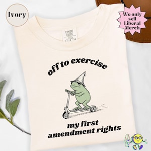 Può includere: Una t-shirt color avorio con la grafica di una rana verde che indossa un cappello a punta e va su un monopattino. Il testo sopra la rana recita "off to exercise" e il testo sottostante recita "my first amendment rights" in minuscolo.