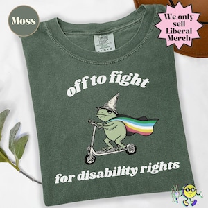 Puede incluir: Una camiseta verde musgo con un gráfico de una rana con sombrero de mago y una capa con los colores de la bandera del orgullo de la discapacidad, montada en un patinete. El texto dice off to fight for disability rights en blanco.