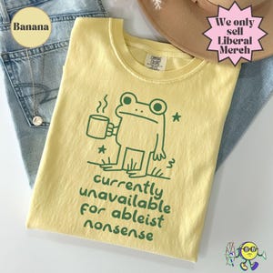 May include: A pale yellow t-shirt featuring a dark green line drawing of a frog holding a steaming mug, with the text "currently unavailable for ableist nonsense" printed below in a rounded green font, and a small Comfort Colors brand tag.