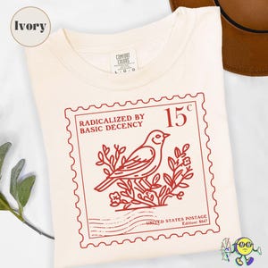 Puede incluir: Camiseta de color marfil con un diseño de sello postal rojo. El sello incluye una ilustración de un pájaro, el texto "RADICALIZED BY BASIC DECENCY" y el precio "15¢." La camiseta es de la marca Comfort Colors, talla L-G-G.