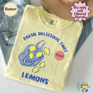 May include: A pale yellow t-shirt featuring a blue graphic of a mesh bag filled with lemons and loose lemons. The text reads FRESH DELICIOUS TART LEMONS and 2547 inside an orange starburst shape. The shirt has a Comfort Colors brand neck label.