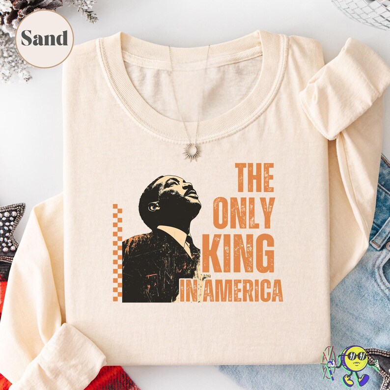 Folded long-sleeve shirt in "Sand" color with a round neckline. Front graphic features a stylized black and orange portrait of a looking-up man in a suit. Orange text beside the image reads "THE ONLY KING IN AMERICA" in bold, distressed block letters. A vertical line of small orange squares frames the left side of the portrait. The shirt is laid flat over denim fabric and a red plaid cloth.