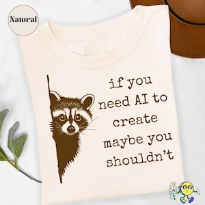 May include: A natural-coloured t-shirt featuring a dark brown graphic of a raccoon peeking from the left side. The text printed in dark brown reads: if you need AI to create maybe you shouldn't. The shirt has a round neckline and short sleeves.