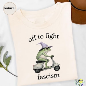 Könnte beinhalten: Cremefarbenes T-Shirt mit dem Text "off to fight fascism". Die Grafik zeigt einen grünen Frosch mit lila Hexenhut, der einen weißen Roller fährt. Das Shirt hat ein "Natural"-Etikett.