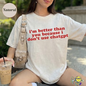 May include: A cream-coloured short-sleeve t-shirt featuring the red text 'i'm better than you because i don't use chatgpt' printed on the front. The shirt has a crew neckline and a relaxed fit, shown with a beige shoulder bag and a plastic cup.