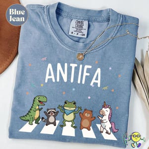 Puede incluir: Camiseta azul vaquero con la palabra "ANTIFA" en letras blancas. Debajo del texto hay un gráfico de animales de dibujos animados cruzando un paso de cebra blanco. La camiseta tiene una etiqueta Comfort Colors y un collar dorado.