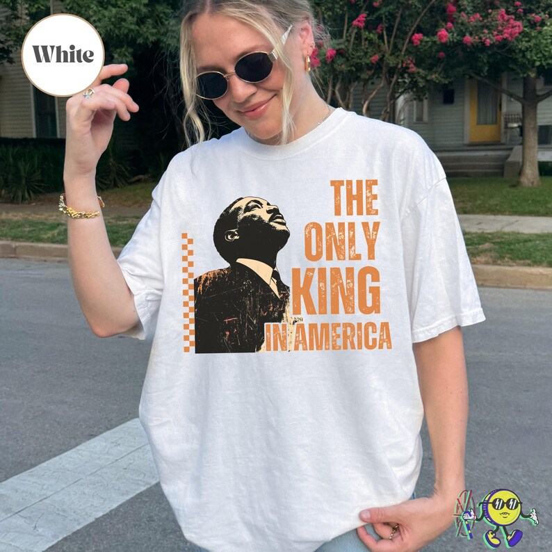 White oversized t-shirt worn by a model, featuring a vintage-style graphic of Martin Luther King Jr. in black and beige with orange accents. Text in bold, distressed orange letters reads "THE ONLY KING IN AMERICA" to the right of the image. A vertical orange checkerboard pattern is on the left side of the graphic.