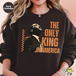 Dark chocolate sweatshirt with a vintage-style graphic of a man looking upward in black and cream tones. To the right, distressed large orange text reads "THE ONLY KING IN AMERICA." A vertical orange checkerboard stripe borders the left side of the image. A model wears the sweatshirt, with the focus on the chest area.