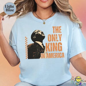 Light blue short-sleeve T-shirt with a black and orange stylized portrait of a man looking upward. Orange text on the right reads "THE ONLY KING IN AMERICA" in bold, distressed font. Orange checkerboard vertical line detail to the left of the portrait. The shirt has a relaxed fit and worn by a smiling model.