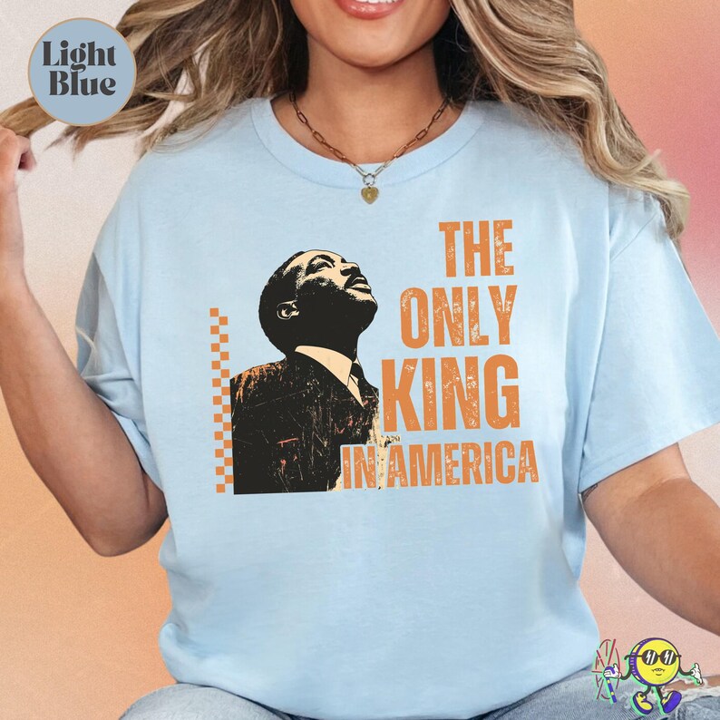 Light blue short-sleeve T-shirt with a black and orange stylized portrait of a man looking upward. Orange text on the right reads "THE ONLY KING IN AMERICA" in bold, distressed font. Orange checkerboard vertical line detail to the left of the portrait. The shirt has a relaxed fit and worn by a smiling model.
