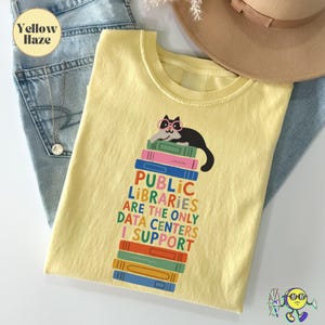 Op de afbeelding: Een lichtgeel t-shirt met een afbeelding van een zwarte kat met een roze bril die op een stapel kleurrijke boeken ligt. De tekst luidt: PUBLIC LIBRARIES ARE THE ONLY DATA CENTERS I SUPPORT. Het t-shirt ligt op een blauwe spijkerstof.