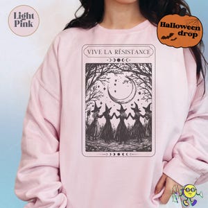 Light pink sweatshirt featuring a black illustration of five witches holding hands in a forest clearing under a crescent moon and stars. Above the image is the text "VIVE LA RÉSISTANCE" in a framed box. The design includes moon phase symbols above and below the illustration, creating a mystical, Halloween-themed vibe.