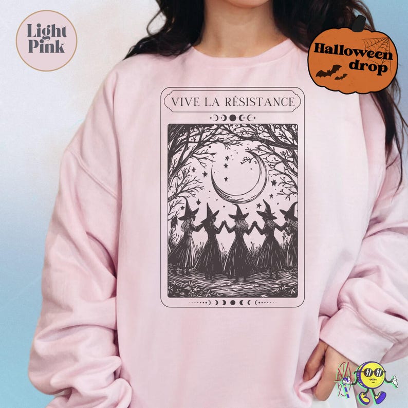 Light pink sweatshirt featuring a black illustration of five witches holding hands in a forest clearing under a crescent moon and stars. Above the image is the text "VIVE LA RÉSISTANCE" in a framed box. The design includes moon phase symbols above and below the illustration, creating a mystical, Halloween-themed vibe.