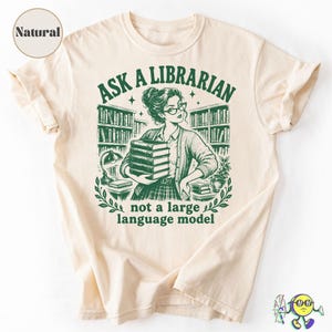 Może przedstawiać: T-shirt w naturalnym kolorze z ciemnozieloną grafiką przedstawiającą osobę w okularach trzymającą stos książek w bibliotece. Tekst brzmi ASK A LIBRARIAN not a large language model. Wzór zawiera regały z książkami, lampę oraz rośliny.
