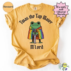 Può includere: T-shirt color senape con un disegno di una rana con un mantello arcobaleno e una spada. Il testo sulla maglietta recita "Twas the Tap Water M'Lord" in carattere gotico nero. Una stella rosa dice "We only sell Liberal Merch".