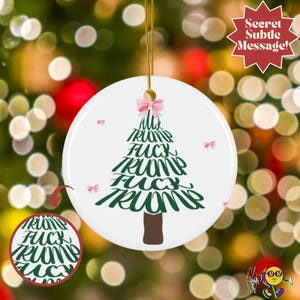 Può includere: Ornamento natalizio rotondo in ceramica bianca con un disegno di albero di Natale verde formato dalle parole "FUCK TRUMP". Un fiocco rosa adorna la parte superiore. L'ornamento è sospeso da un filo d'oro. Sfondo sfocato di luci natalizie.