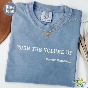 May include: A blue jean colored t-shirt with the text "TURN THE VOLUME UP - Mayor Mamdani" in white. A gold necklace with a circular pendant rests on the shirt. The shirt is folded and has a Comfort Colors tag.