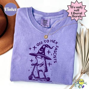 Op de afbeelding: Een violetkleurig T-shirt met korte mouwen en een donkerpaarse afbeelding van een kikker met een heksenhoed met sterren op een step. De tekst luidt OFF TO HEX FASCISTS. Het T-shirt is voorzien van een Comfort Colors label en ronde hals.