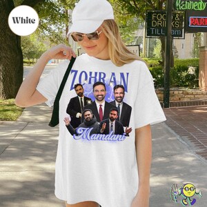 White oversized T-shirt featuring five different images of a man with dark hair and beard, wearing suits and casual clothes, arranged in a collage. Above images, text reads "ZOHRAN" in large bold purple letters with lightning effects behind. Below, the name "Mamdani" is written in elegant, blue cursive font. A model wears the T-shirt with a white cap and sunglasses.