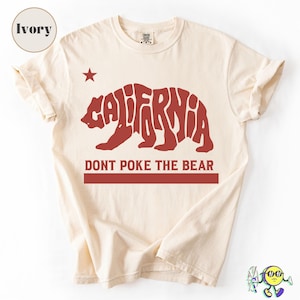 May include: Ivory-colored t-shirt featuring a red bear-shaped design with the word "CALIFORNIA" and the phrase "DONT POKE THE BEAR." A red star is above the bear. The shirt is made of a soft material.