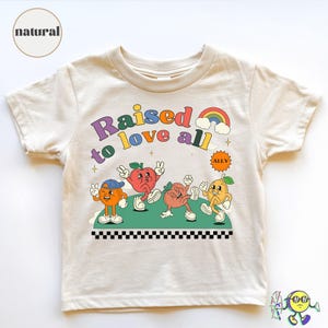 May include: Cream-coloured t-shirt with the text "Raised to love all" in colourful retro font. The shirt features cartoon fruit characters, a rainbow, and the word "ALLY". The shirt has a black and white chequered border.