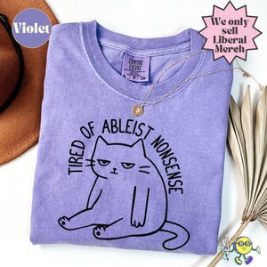 May include: A violet short-sleeved t-shirt featuring a black line drawing of a cat sitting down with the text "TIRED OF ABLEIST NONSENSE" arched above it. The shirt has a crew neckline and a Comfort Colors brand label on the inside collar.