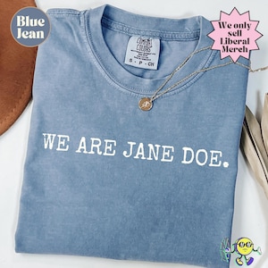 以下が含まれることがあります： フロントに白い文字で「WE ARE JANE DOE.」とプリントされたブルーデニムカラーのTシャツ。首元にはComfort Colorsのブランドラベルが付いており、小さな円形ペンダントが付いたゴールドトーンのネックレスが添えられています。