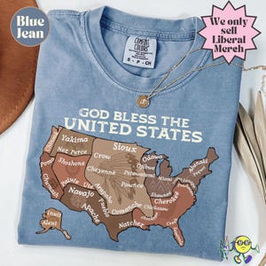 Puede incluir: Camiseta azul vaquero con el texto "GOD BLESS THE UNITED STATES" y un mapa de EE. UU. con nombres de tribus. La camiseta también tiene el texto "We only sell Liberal Merch". Un collar dorado está en la camiseta.