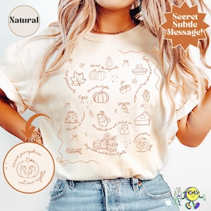 May include: Cream-colored t-shirt with a fall-themed graphic. The design includes pumpkins, leaves, pie, and text that reads "carve pumpkins not our rights". The shirt has a round neckline and short sleeves.