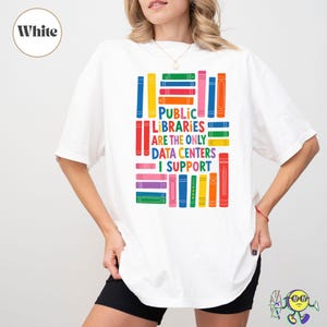May include: A white short-sleeved t-shirt featuring a colorful graphic of stacked books and the text "PUBLIC LIBRARIES ARE THE ONLY DATA CENTERS I SUPPORT" printed in the center. The design uses various bright colors including red, blue, green, and yellow.