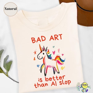 Może przedstawiać: T-shirt w naturalnym kolorze z kolorowym rysunkiem jednorożca w stylu kredkowym. Nad jednorożcem znajduje się czerwony napis "BAD ART", a pod nim czerwony napis "is better than AI slop". Wzór zawiera również małe gwiazdki i serduszka.