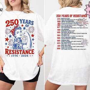 May include: A white t-shirt featuring a red, white, and blue graphic of a person flexing with the text 250 YEARS OF RESISTANCE 1776-2026. The back displays a timeline of historical events from 1773 to 2020 with red and blue firework illustrations.