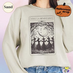 Sand-colored sweatshirt with a black vintage-style graphic of five witches holding hands in a forest clearing under a crescent moon and stars, framed by bare tree branches. Above the image is the text "VIVE LA RÉSISTANCE" with phases of the moon illustrated above and below the graphic. A model wears the sweatshirt paired with blue jeans. A small pumpkin graphic says "Halloween drop."