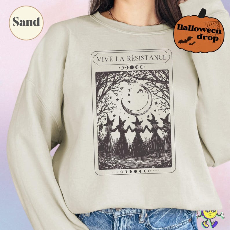 Sand-colored sweatshirt with a black vintage-style graphic of five witches holding hands in a forest clearing under a crescent moon and stars, framed by bare tree branches. Above the image is the text "VIVE LA RÉSISTANCE" with phases of the moon illustrated above and below the graphic. A model wears the sweatshirt paired with blue jeans. A small pumpkin graphic says "Halloween drop."