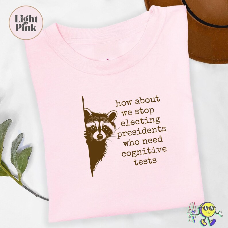 Light pink folded t-shirt with a brown graphic of a raccoon peeking from behind a line and the text “how about we stop electing presidents who need cognitive tests” in typewriter-style font. A brown hat and a green leafy branch are partially visible around the shirt. A circular label in the top left reads “Light Pink.”