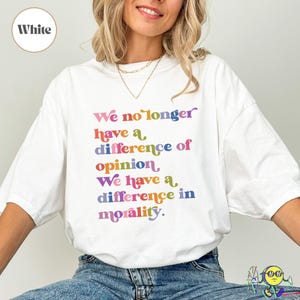 Peut inclure: T-shirt blanc avec l'inscription "We no longer have a difference of opinion, We have a difference in morality" en lettres colorées de style rétro. Le t-shirt a un col rond et des manches courtes.