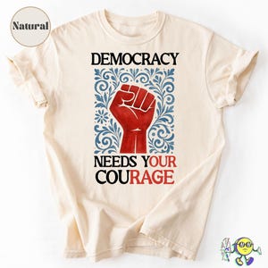 May include: Cream-colored t-shirt with the words "DEMOCRACY NEEDS YOUR COURAGE" in black and red. A red fist graphic is centered within a blue and white floral design. The shirt has short sleeves and a relaxed fit.