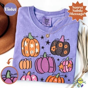 Può includere: Una t-shirt viola a maniche corte con una stampa grafica di varie zucche in arancione, nero e rosa, decorate con stelle e fiori. La maglietta ha un'etichetta Comfort Colors sul collo e include del testo sulle zucche.