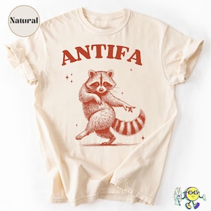 Puede incluir: Camiseta color crema con la palabra "ANTIFA" en letras rojas en bloque sobre una ilustración de un mapache bailando. El mapache es rojo con una cola a rayas y está bailando con un brazo levantado. La camiseta tiene una etiqueta "Natural".