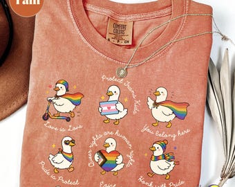 May include: Coral Comfort Colors t-shirt with cartoon ducks and LGBTQ+ pride symbols. Text includes "Love is Love", "Pride is Protest", "Queer rights are human rights", "You belong here", and "Honk with Pride". A silver necklace is draped across the shirt.